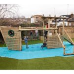 Children are playing on a wooden playground structure designed like a ship, with climbing nets and slides, on artificial grass and blue safety surfacing.