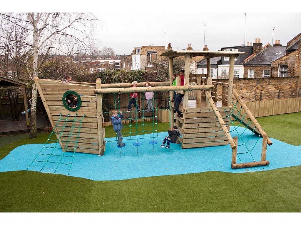 Children are playing on a wooden playground structure designed like a ship, with climbing nets and slides, on artificial grass and blue safety surfacing.