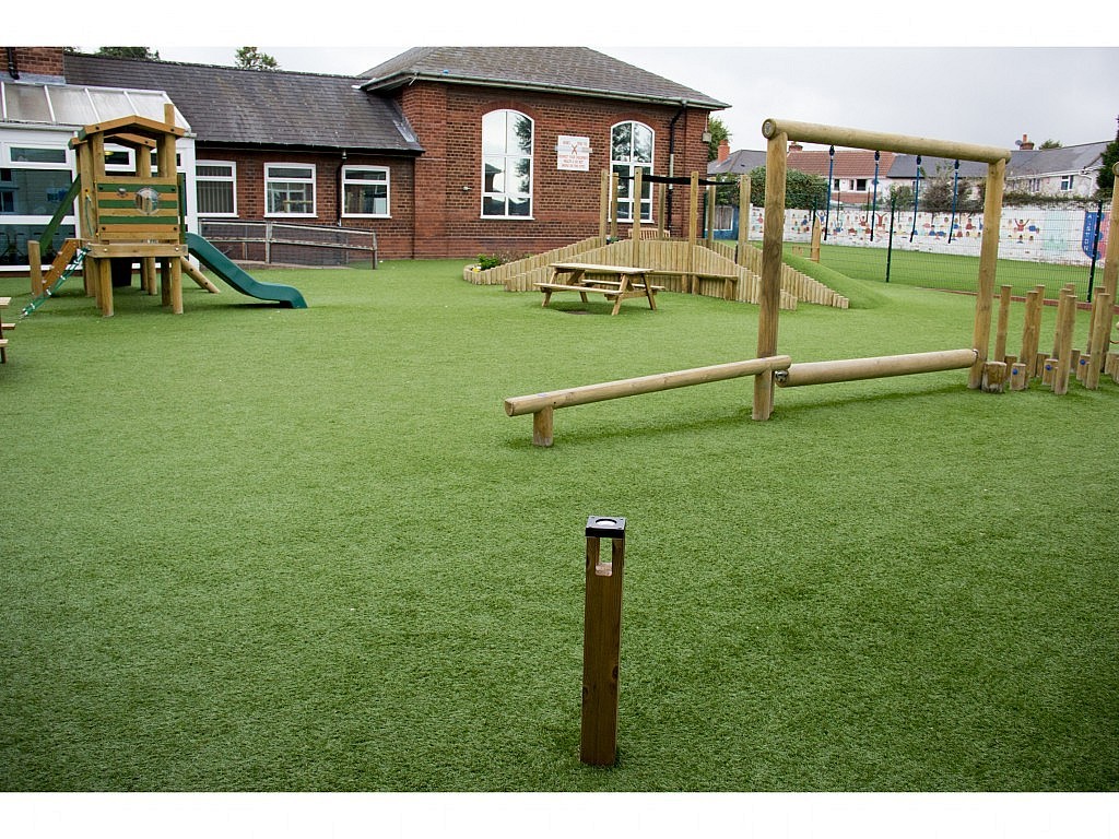 A school playground with artificial grass, a climbing frame with a slide, wooden play equipment, picnic tables, and a brick school building in the background.