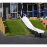 A small metal slide descends from a grassy artificial hill with wooden sides in an outdoor playground area next to a building.