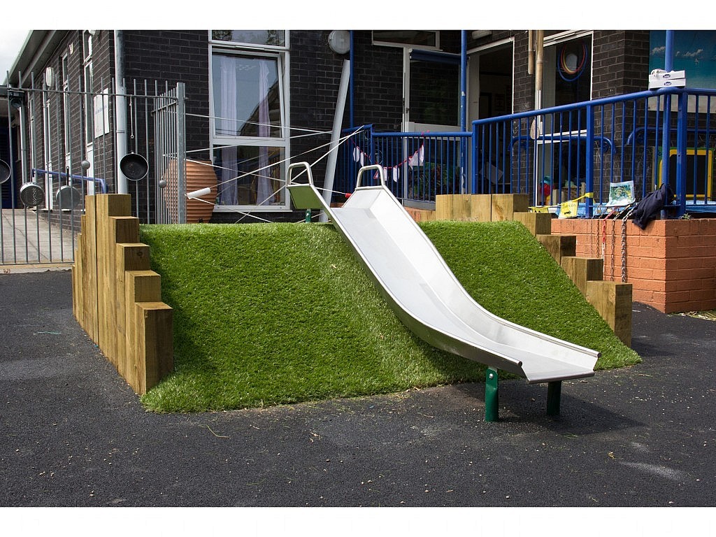 A small metal slide descends from a grassy artificial hill with wooden sides in an outdoor playground area next to a building.