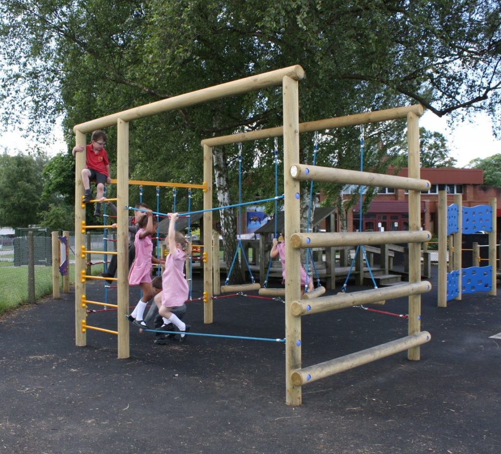 Children in school uniforms play on a wooden climbing frame and rope structure at a playground, surrounded by trees and outdoor buildings.
