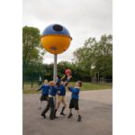 Four children in school uniforms play with a red ball under a large, colourful spherical playground structure mounted on a pole. Trees and a fenced area are visible in the background.