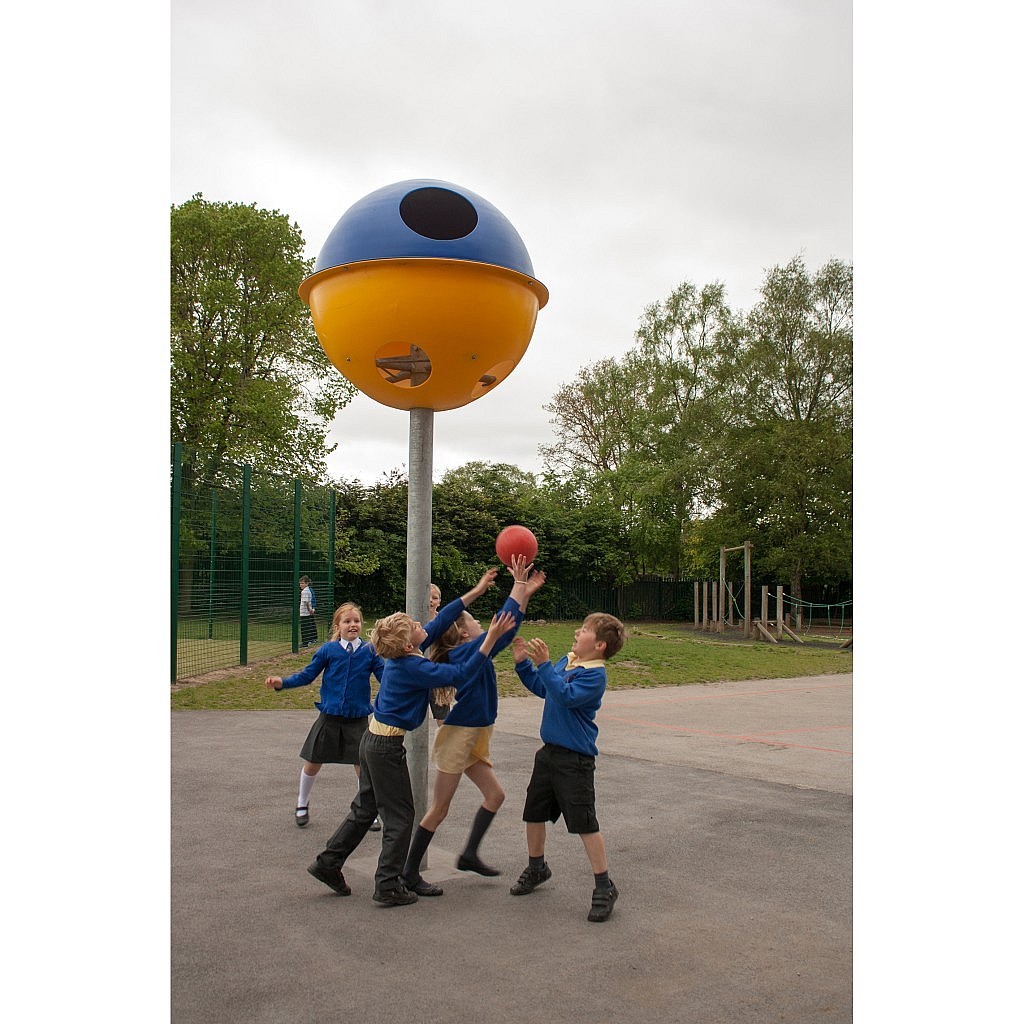 Four children in school uniforms play with a red ball under a large, colourful spherical playground structure mounted on a pole. Trees and a fenced area are visible in the background.