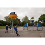 Children in school uniforms play near a large yellow and blue playground structure and a fenced basketball hoop on an outdoor court surrounded by trees.