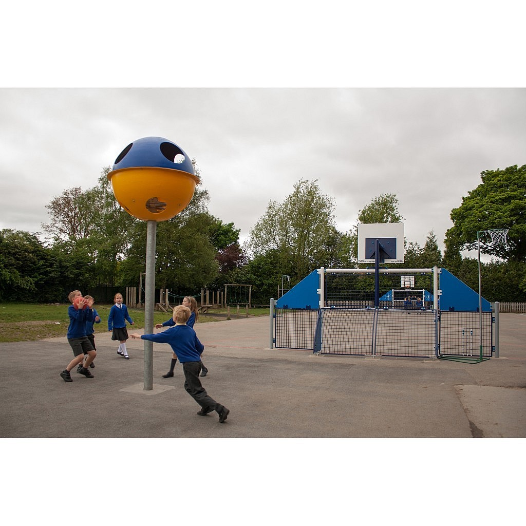 Children in school uniforms play near a large yellow and blue playground structure and a fenced basketball hoop on an outdoor court surrounded by trees.