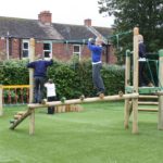 Four children are playing on a wooden playground structure with balance beams and ropes in a grassy glade near residential houses.