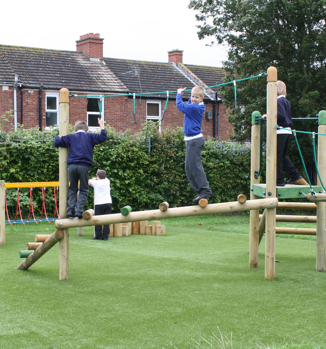 Four children are playing on a wooden playground structure with balance beams and ropes in a grassy glade near residential houses.
