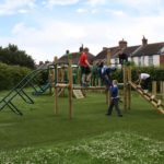 Several children play and climb on a wooden playground structure with slides and bridges in a grassy glade near houses.