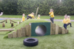 Six young children in yellow jumpers play on and around a grassy hummock hill mound with a tunnel and wooden climbing structures in an outdoor playground.