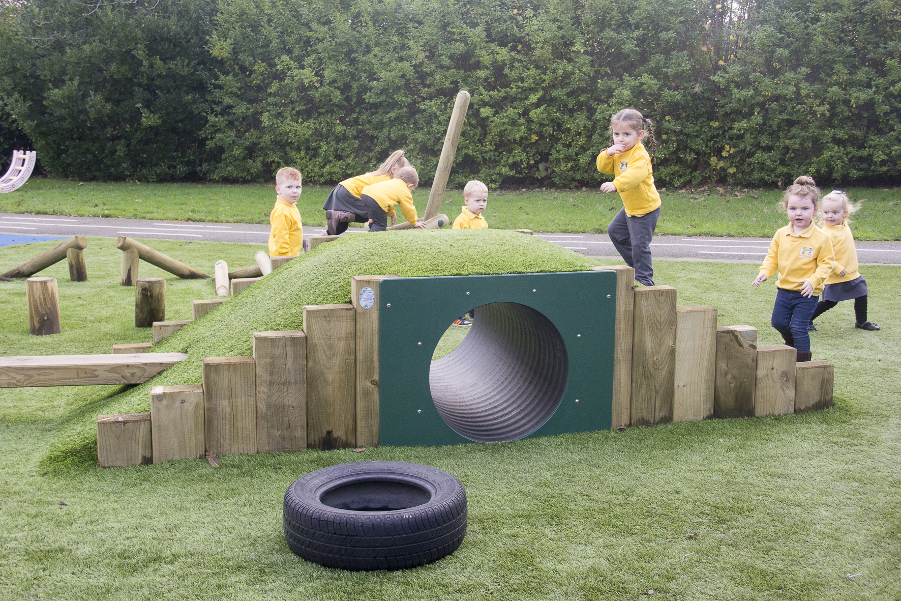 Six young children in yellow jumpers play on and around a grassy hummock hill mound with a tunnel and wooden climbing structures in an outdoor playground.