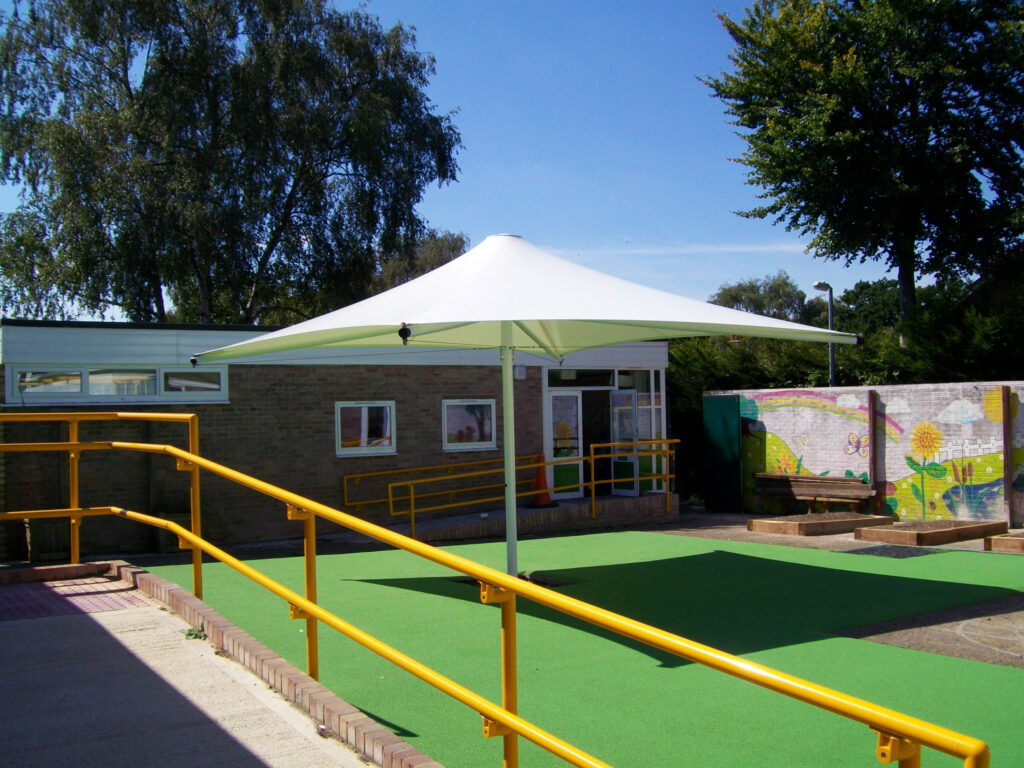 A school courtyard with green artificial grass, a white canopy, yellow railings, and a mural on a brick wall under a clear blue sky.
