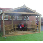 Three children sit on a wooden bench under a covered wooden shelter on artificial grass near a brick building.