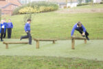 Two boys in school uniforms jump over low hurdles made from wooden beams on a grassy playground while three other children watch from the side.