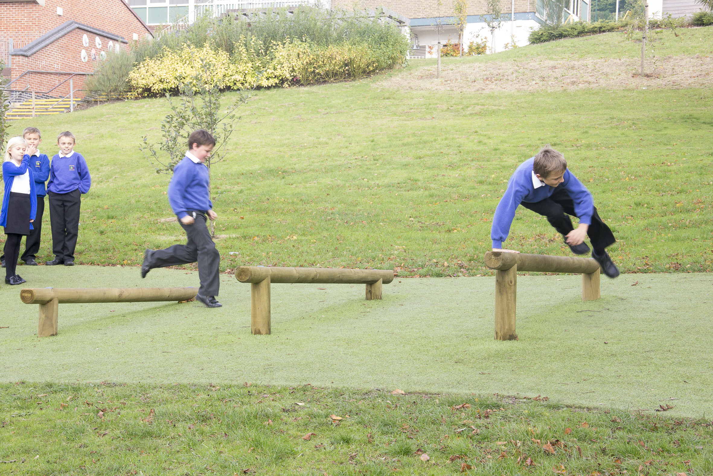 Two boys in school uniforms jump over low hurdles made from wooden beams on a grassy playground while three other children watch from the side.