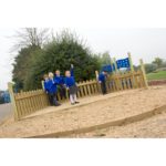 Five children in blue school uniforms stand and play inside a wooden fenced area on a playground, with trees and play equipment in the background.