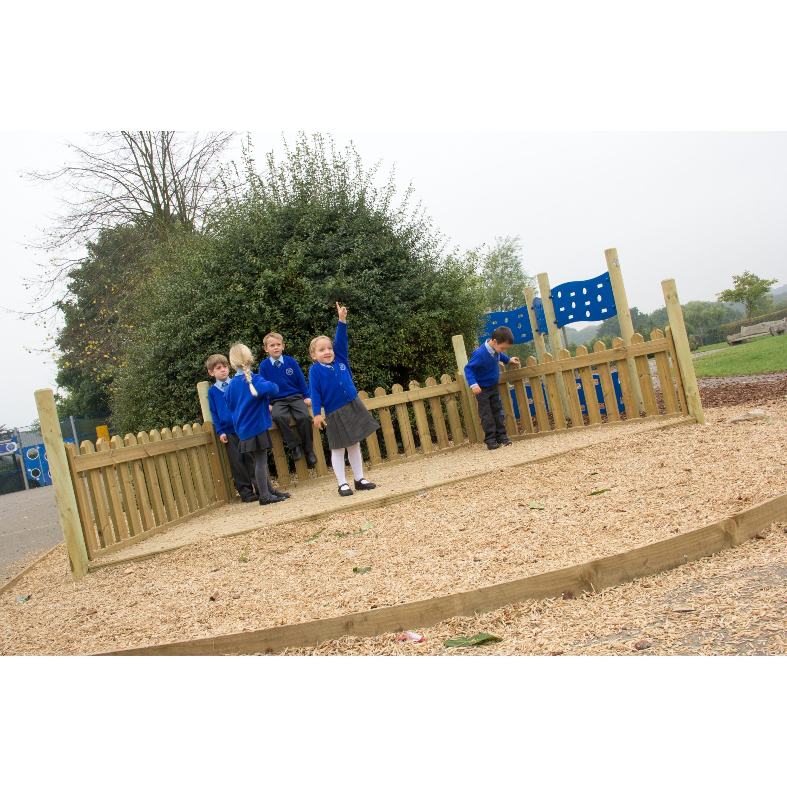 Five children in blue school uniforms stand and play inside a wooden fenced area on a playground, with trees and play equipment in the background.