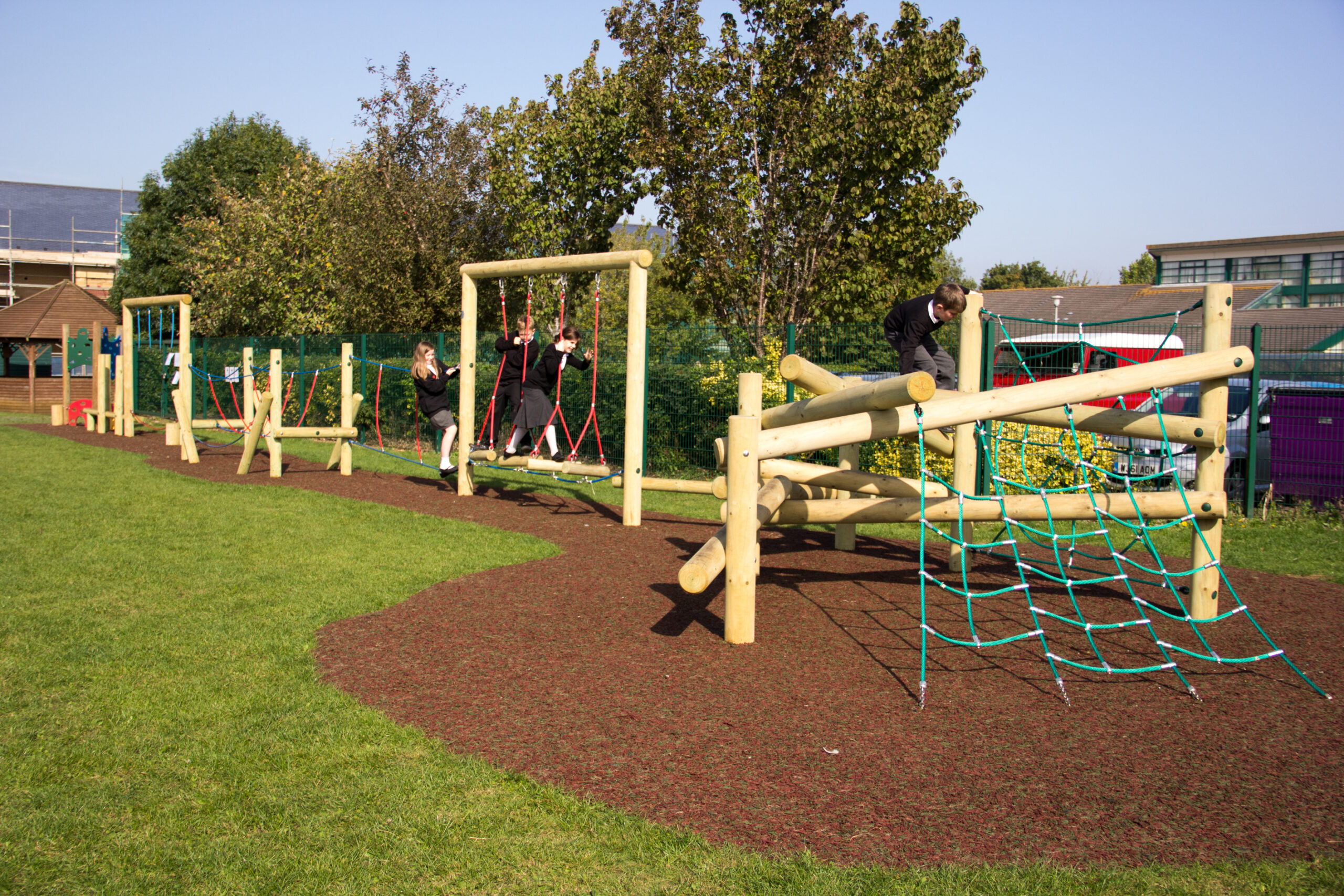 Children play on a wooden Durabond outdoor climbing frame with nets and ropes in a grassy playground area surrounded by trees and a fence.