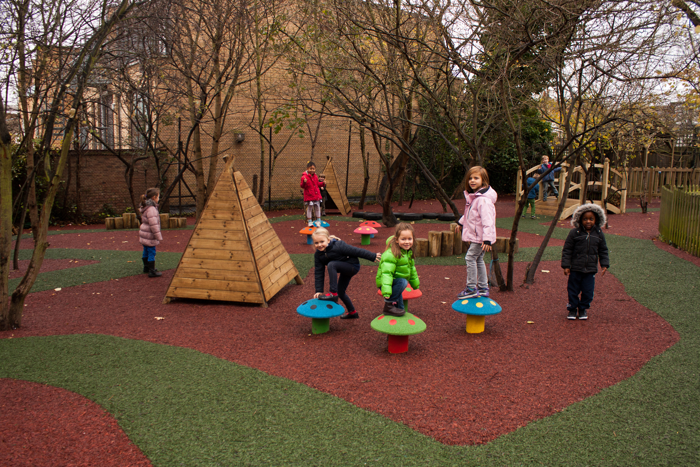 Several children play on colourful, Durabond mushroom-shaped stools and climbing structures in an outdoor playground with artificial grass and trees.