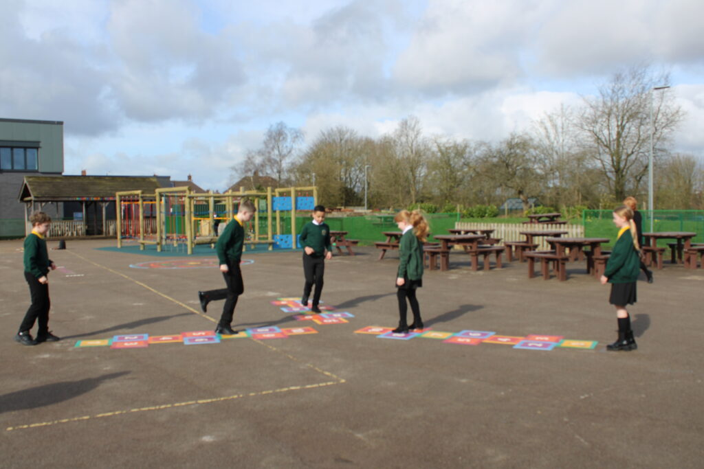 Five children in school uniforms play on a colourful hopscotch grid in a school playground with picnic benches and play structures in the background.