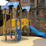 A colourful playground structure featuring the Intrepid 900mm with Poly Deck Slide, climbing nets, and steps, set on a soft surface next to a brick wall and wooden fence.