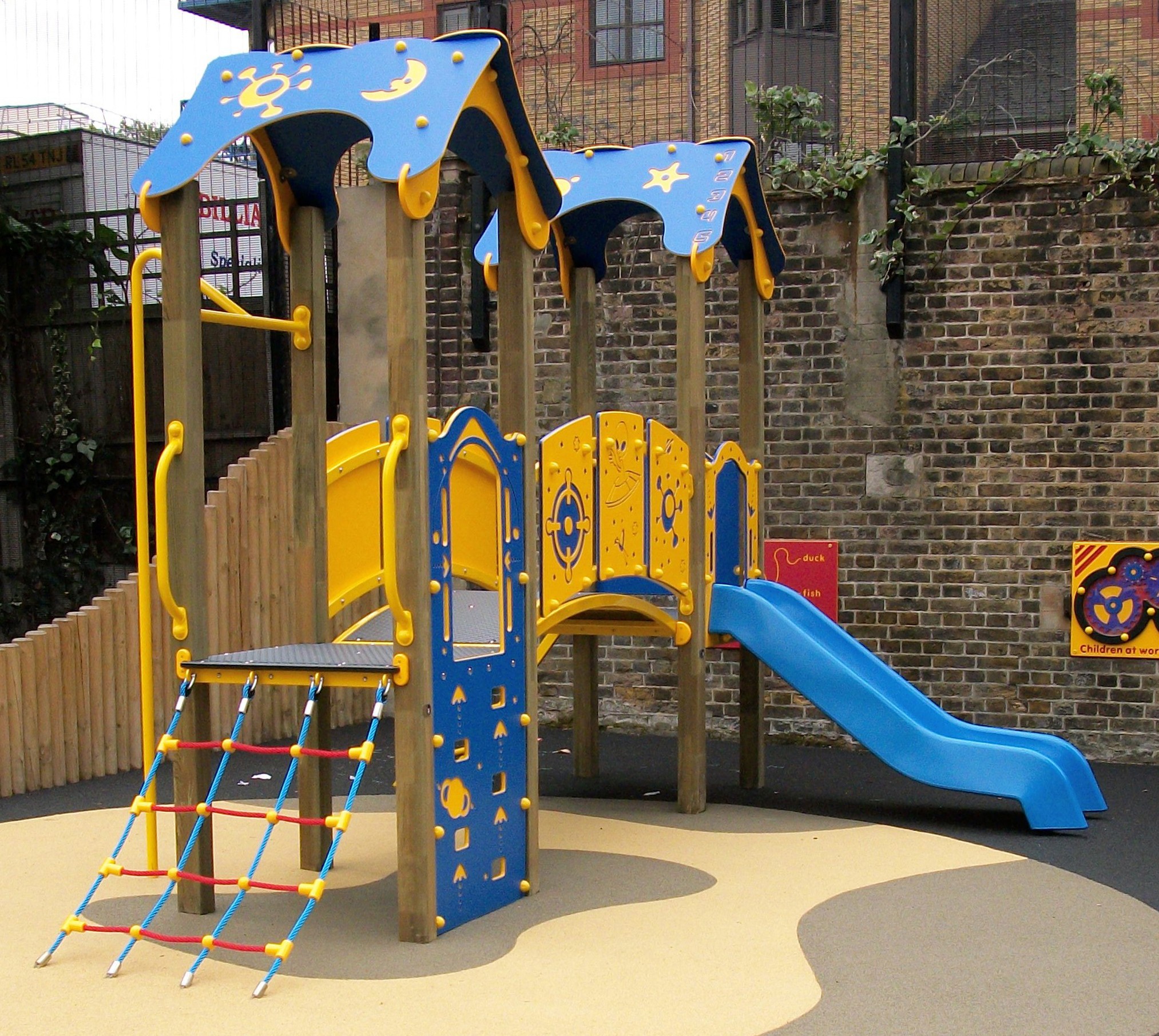 A colourful playground structure featuring the Intrepid 900mm with Poly Deck Slide, climbing nets, and steps, set on a soft surface next to a brick wall and wooden fence.