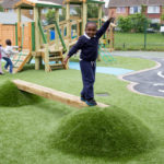 A boy balances on a wooden beam between grassy mounds in a playground, whilst other children play in the background.
