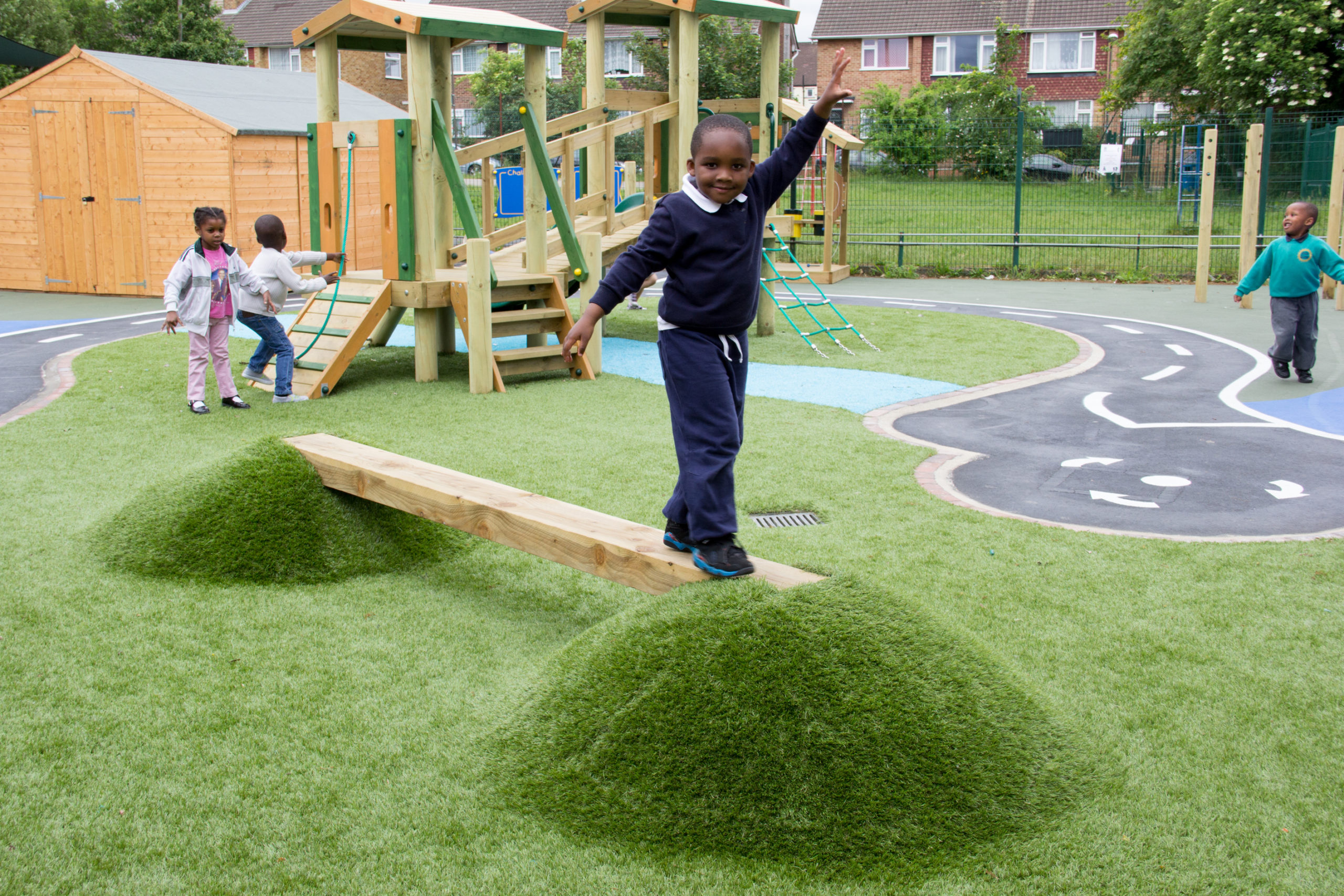 A boy balances on a wooden beam between grassy mounds in a playground, whilst other children play in the background.