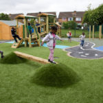 Children play on artificial grass mounds and balance beams in a playground with climbing frames and painted paths, surrounded by wooden fences and houses.