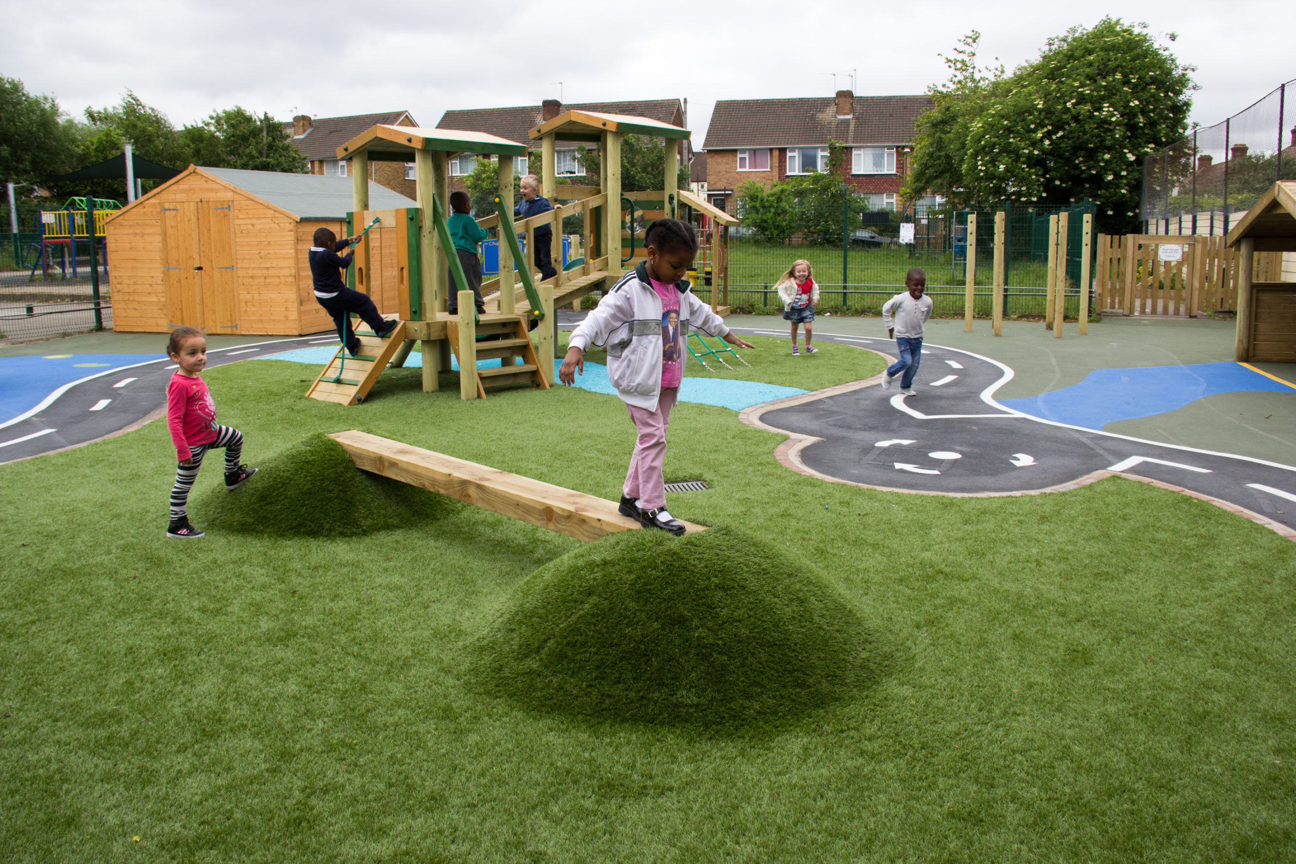 Children play on artificial grass mounds and balance beams in a playground with climbing frames and painted paths, surrounded by wooden fences and houses.