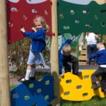 Three young children climb on a colourful Jigsaw Traverse Wall playground structure outdoors, whilst an adult stands in the background.