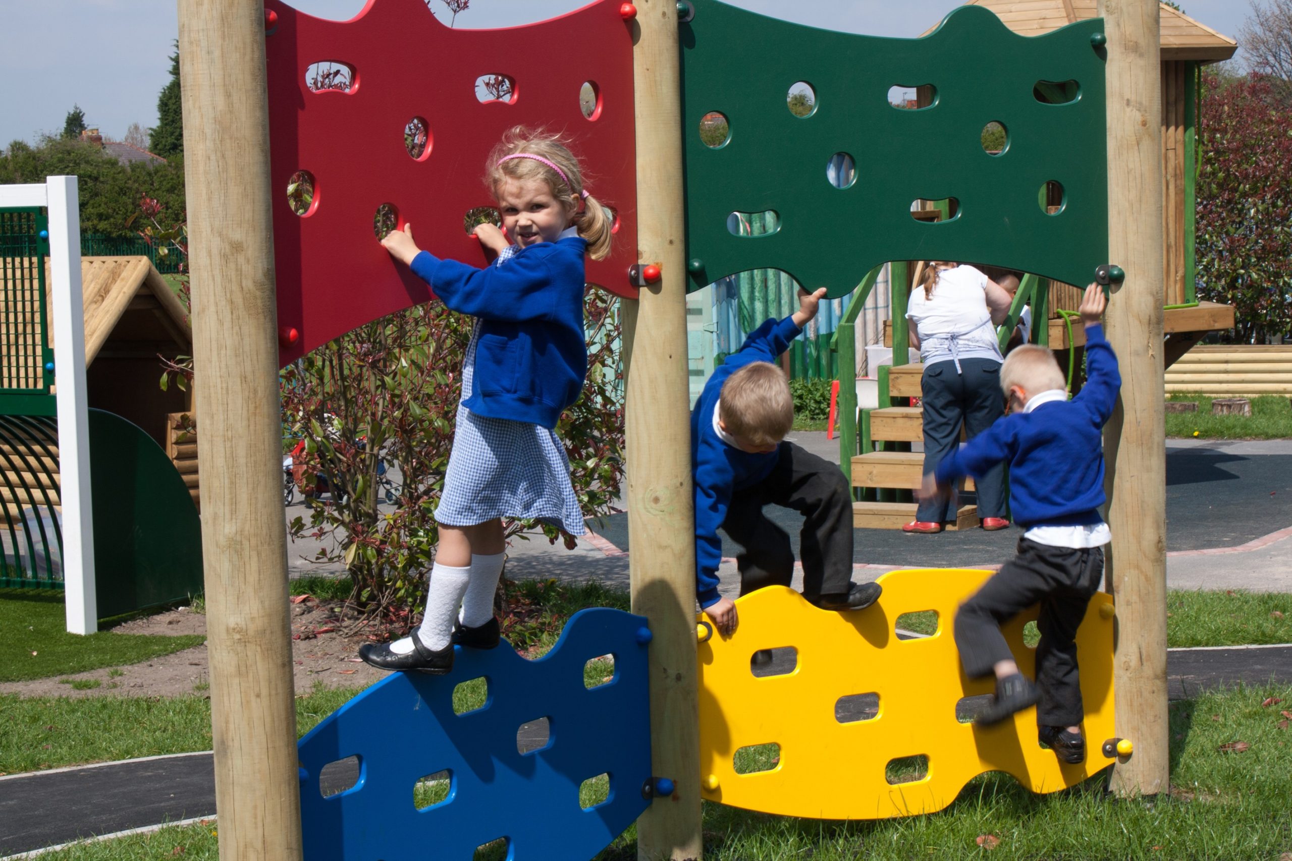 Three young children climb on a colourful Jigsaw Traverse Wall playground structure outdoors, whilst an adult stands in the background.