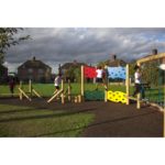Four children play on a wooden playground featuring a Jigsaw Traverse Wall, climbing walls, and rope bridges in an outdoor park with houses and a cloudy sky in the background.