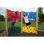 Two boys climb a colourful Jigsaw Traverse Wall with red, blue, green, and yellow panels in an outdoor park setting.