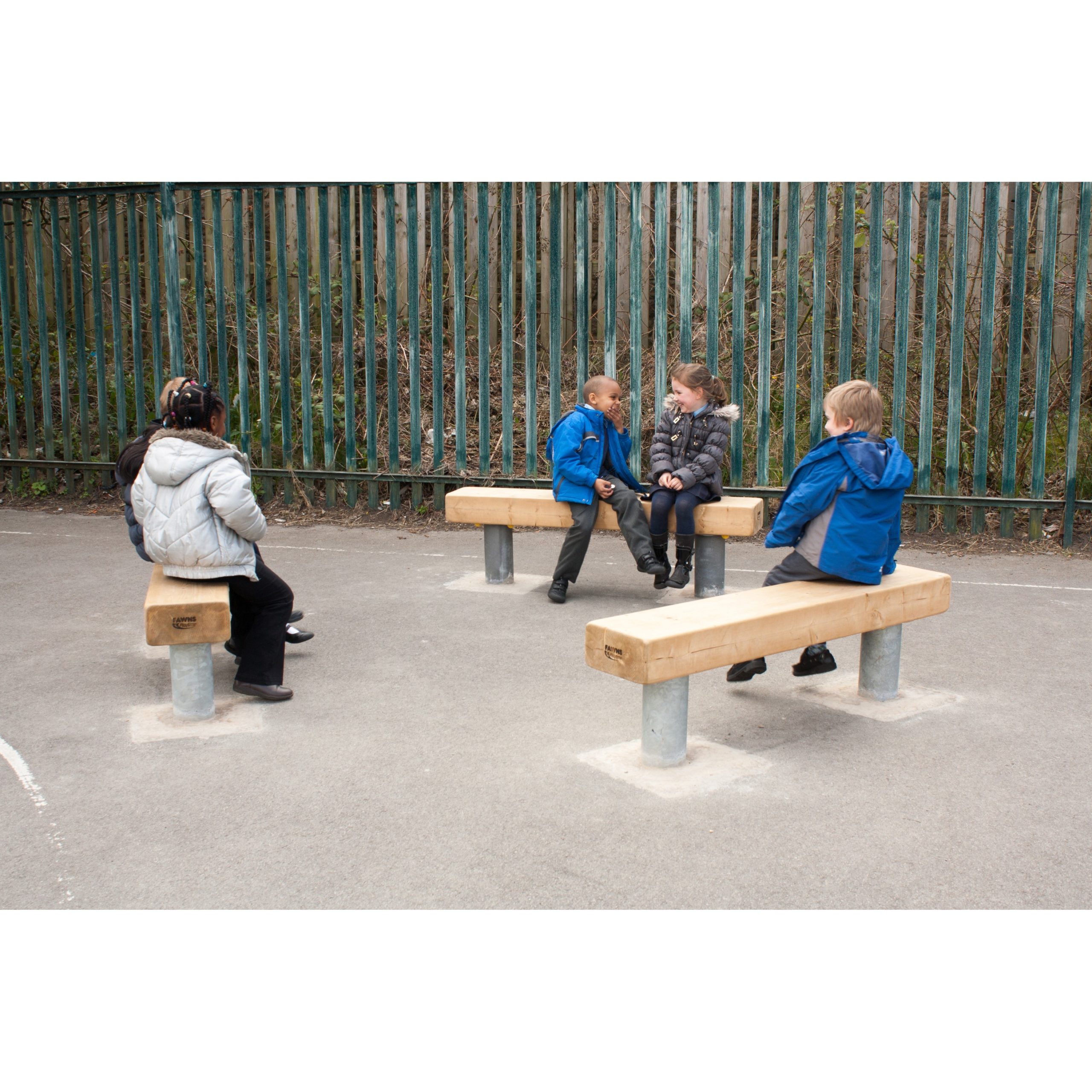 Five children sit on wooden benches arranged in a playground area, with a metal fence and greenery in the background.