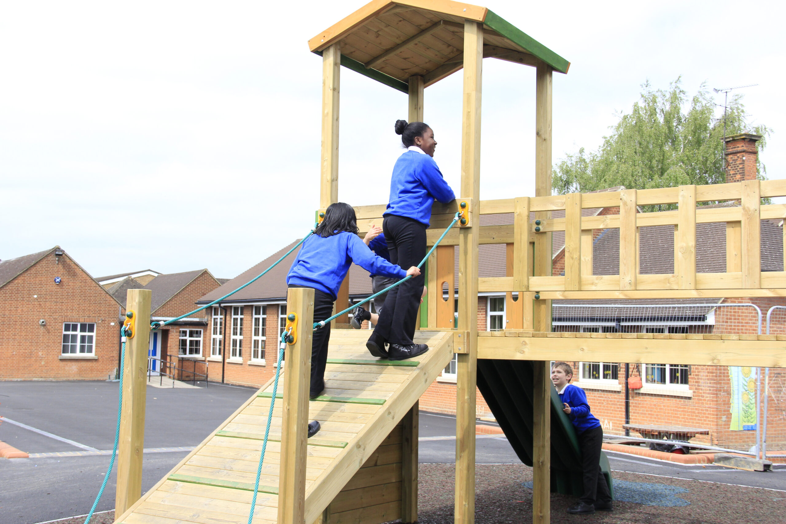 Three young explorers - Cook children in blue jumpers play on a wooden climbing frame in a school playground, with brick buildings in the background.