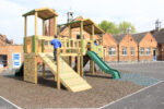 A child in a blue shirt climbs a wooden playground structure with slides and ramps in a schoolyard with brick buildings in the background.