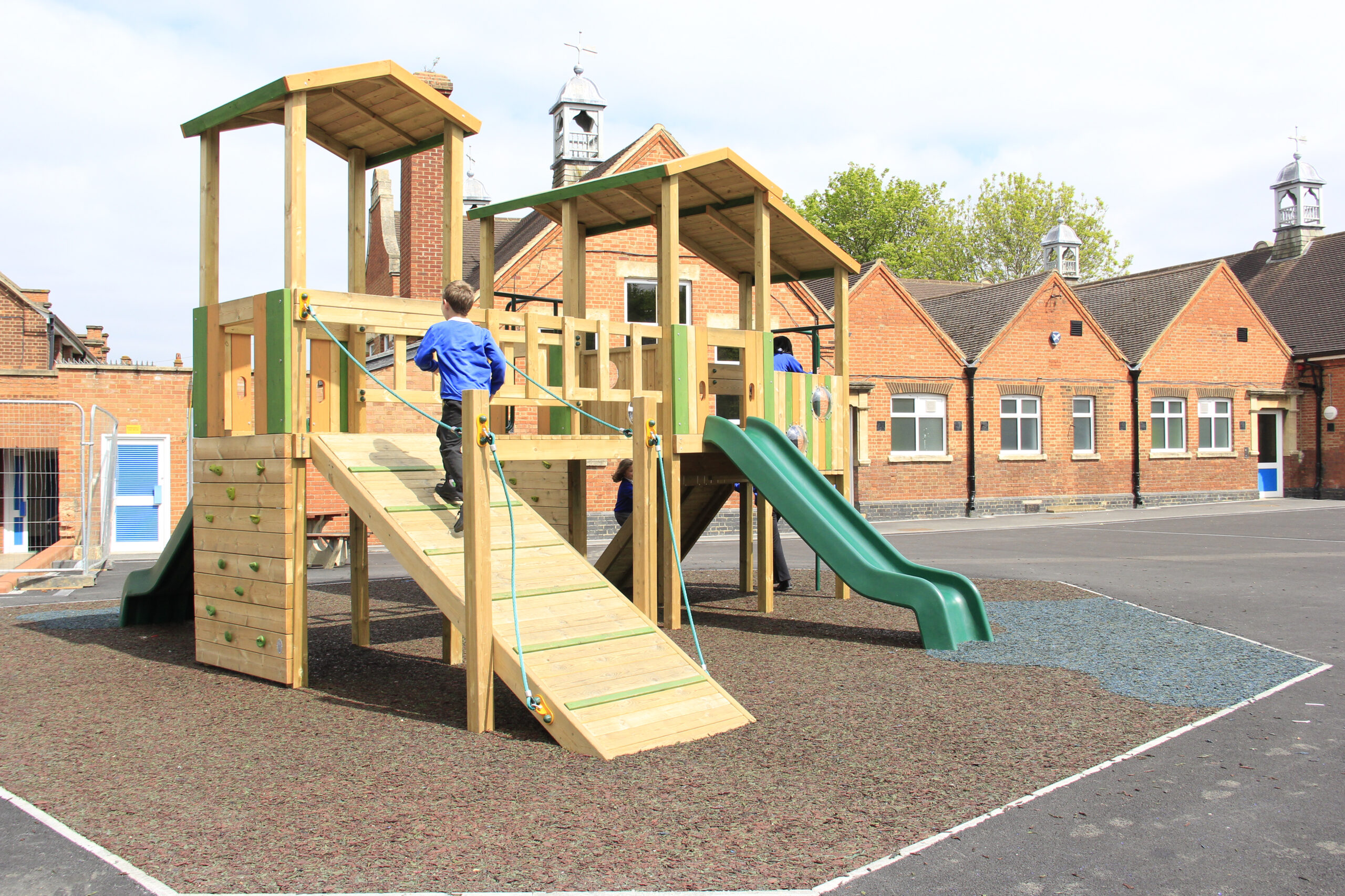A child in a blue shirt climbs a wooden playground structure with slides and ramps in a schoolyard with brick buildings in the background.