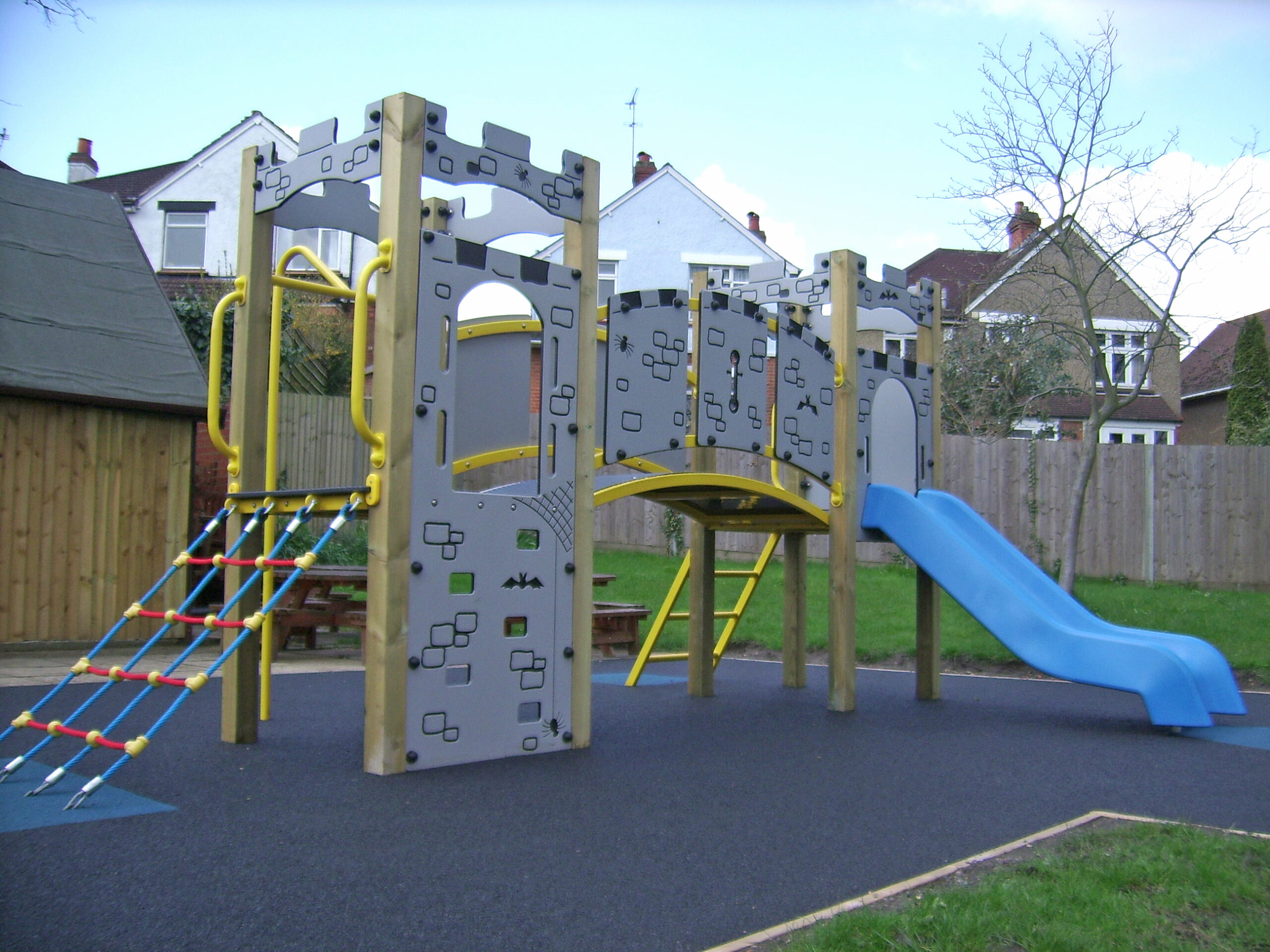 A Lancelot Tower 1200mm with Poly Slide features climbing nets, panels, a bridge, and a blue slide on a rubber surface, set against houses and grass in the background.
