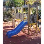 The Lancelot Tower 900mm c/w Poly Slide features a blue double slide, climbing elements, and castle-themed panels on a red rubber surface; trees and a fence are visible in the background.