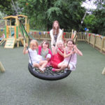Four young children sit together on a round swing while an adult stands behind them in a fenced playground with trees and play equipment in the background.