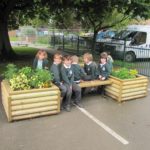 Six children in school uniforms sit and chat on a wooden bench with planter boxes at both ends, enjoying outdoor maths activities using playground equipment under a tree near a fence and parked vehicles.