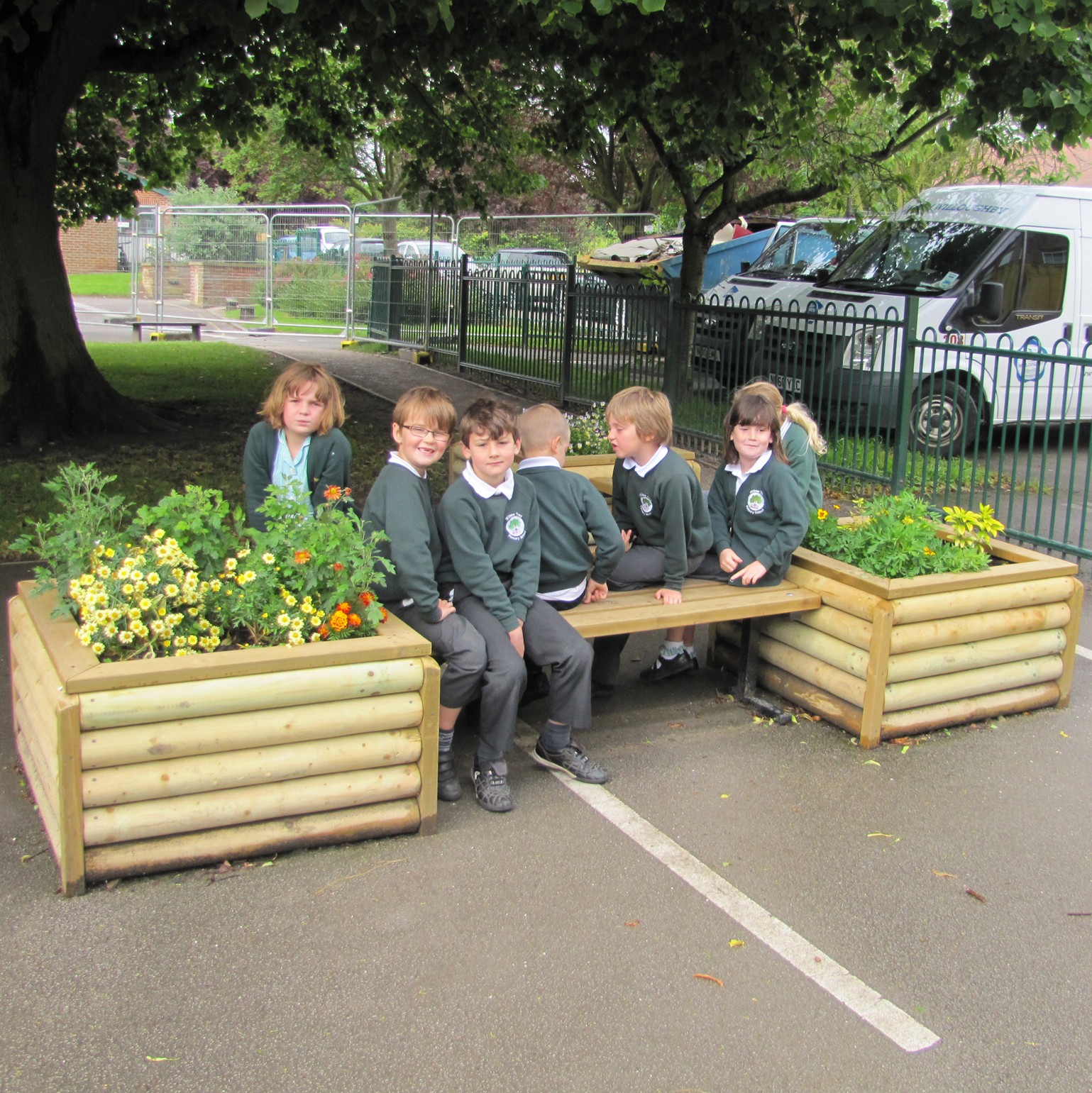 Six children in school uniforms sit and chat on a wooden bench with planter boxes at both ends, enjoying outdoor maths activities using playground equipment under a tree near a fence and parked vehicles.