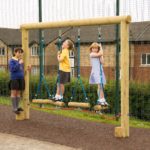 Three children in school uniforms play on a wooden log swing set outdoors, with a fence and houses visible in the background.