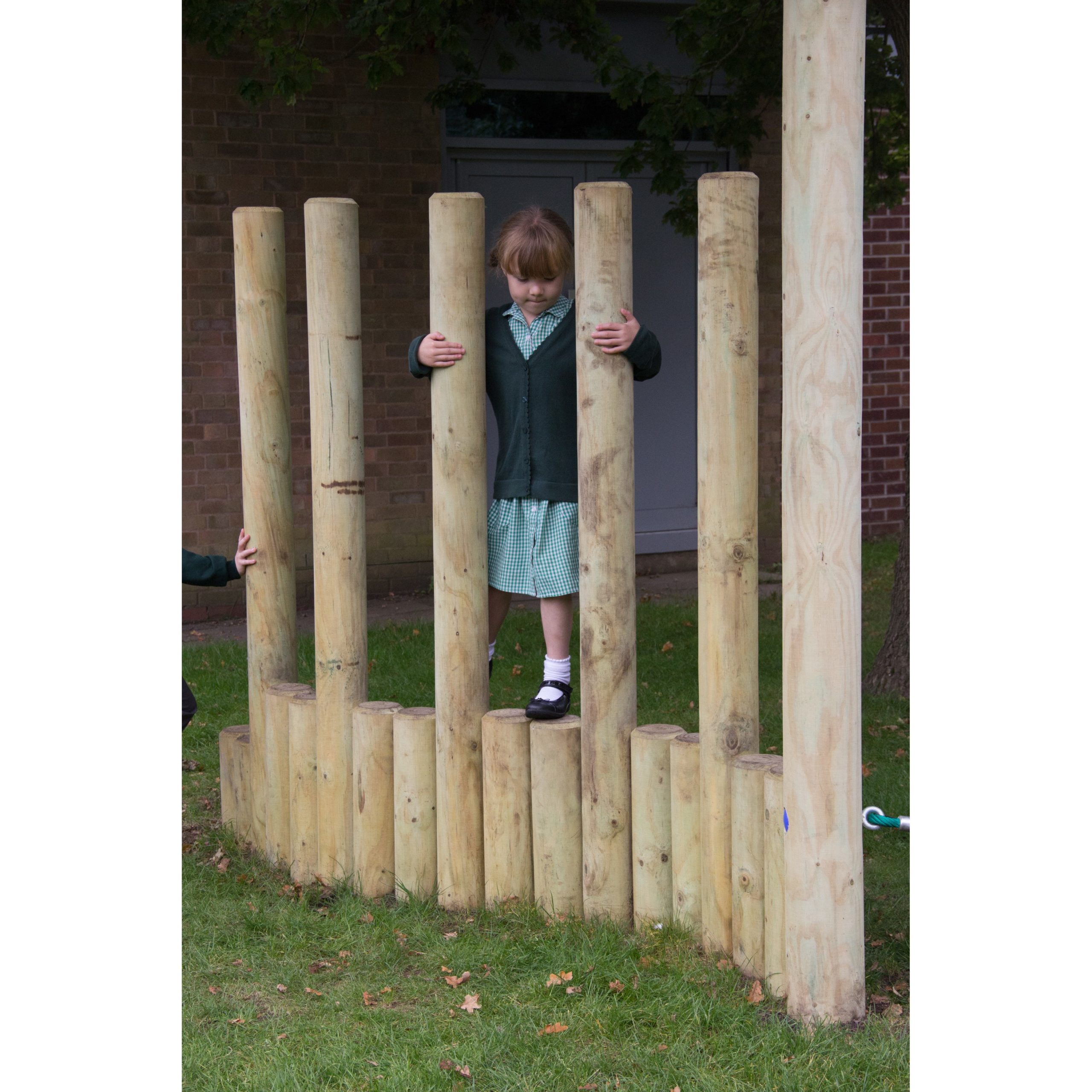 A young girl in a school uniform stands on uneven wooden posts outdoors, holding onto two upright posts for balance.