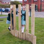 Two children in school uniforms climb and balance on vertical wooden poles in an outdoor playground area.