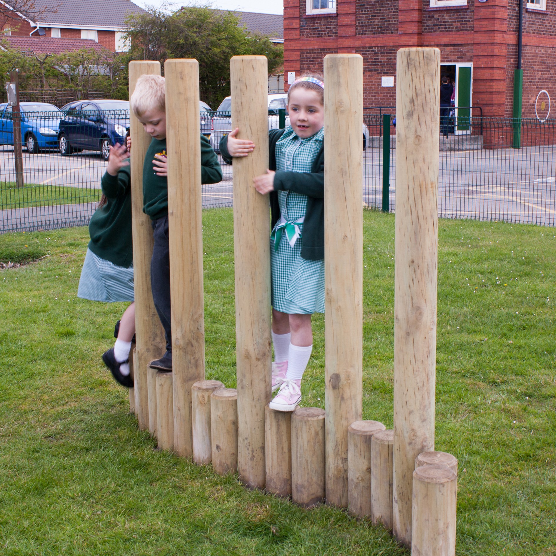 Two children in school uniforms climb and balance on vertical wooden poles in an outdoor playground area.
