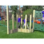 Two children in school uniforms play and climb on vertical wooden poles at an outdoor playground with artificial grass.