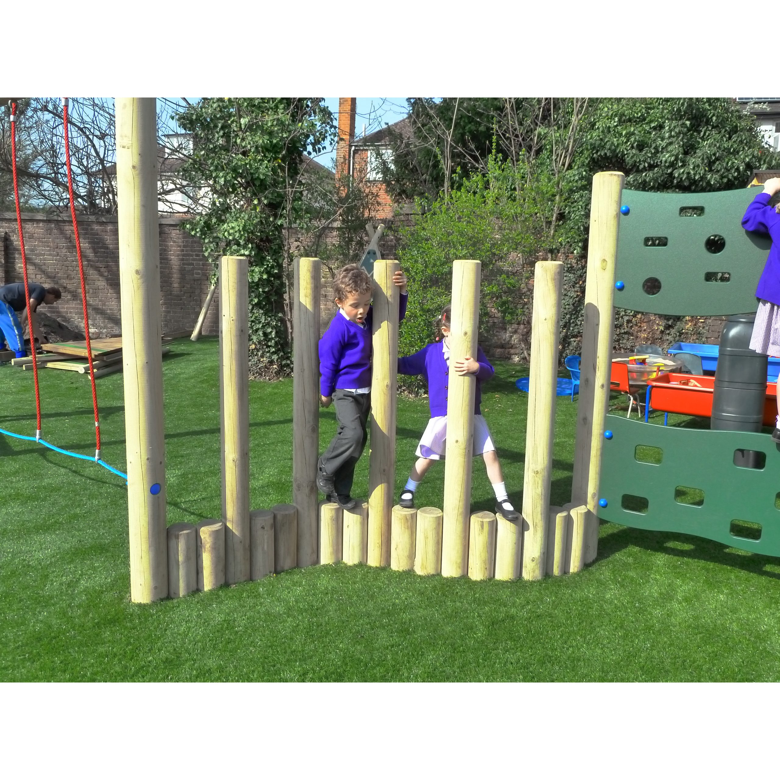 Two children in school uniforms play and climb on vertical wooden poles at an outdoor playground with artificial grass.
