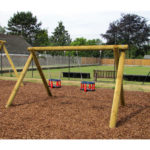 Two empty toddler swings on a wooden frame in a playground with woodchip ground covering and a tennis court in the background.